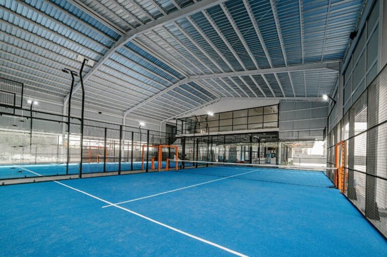 Indoor tennis court with bright blue flooring, surrounded by glass and metal fencing, under a high arched metal roof with natural and artificial lighting.
